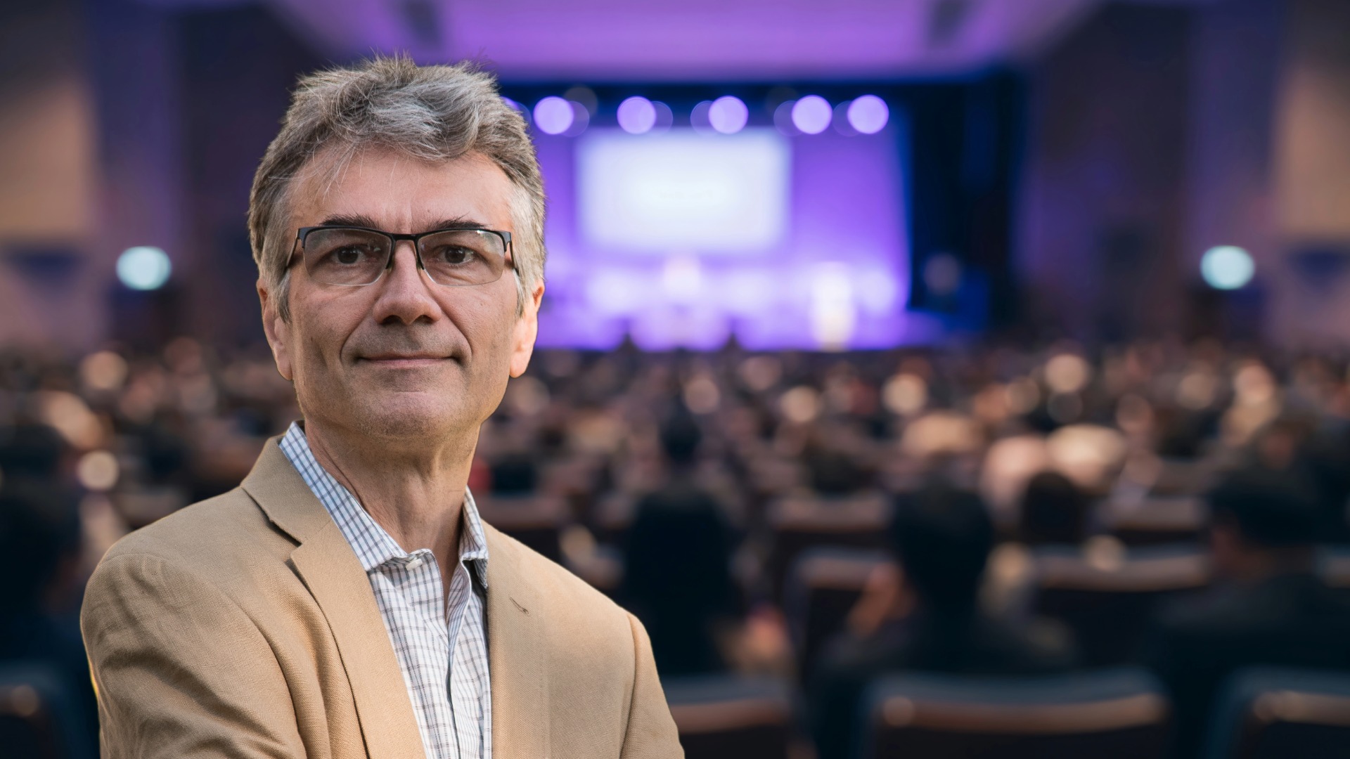 Portrait of Andreas Volmer, Consig's Head of Product, in a tan blazer at a conference, with the audience and stage screen blurred behind him.